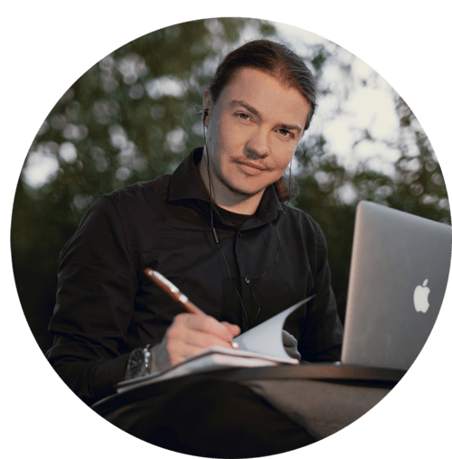 A portrait of Angel Pretot, sitting in nature with his laptop, looking at the camera and using a pen and notebook with his right hand.