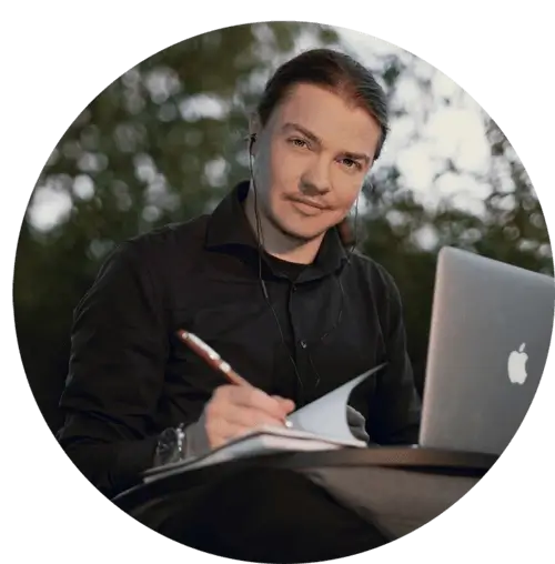 A portrait of Angel Pretot, sitting in nature with his laptop, looking at the camera and using a pen and notebook with his right hand.
